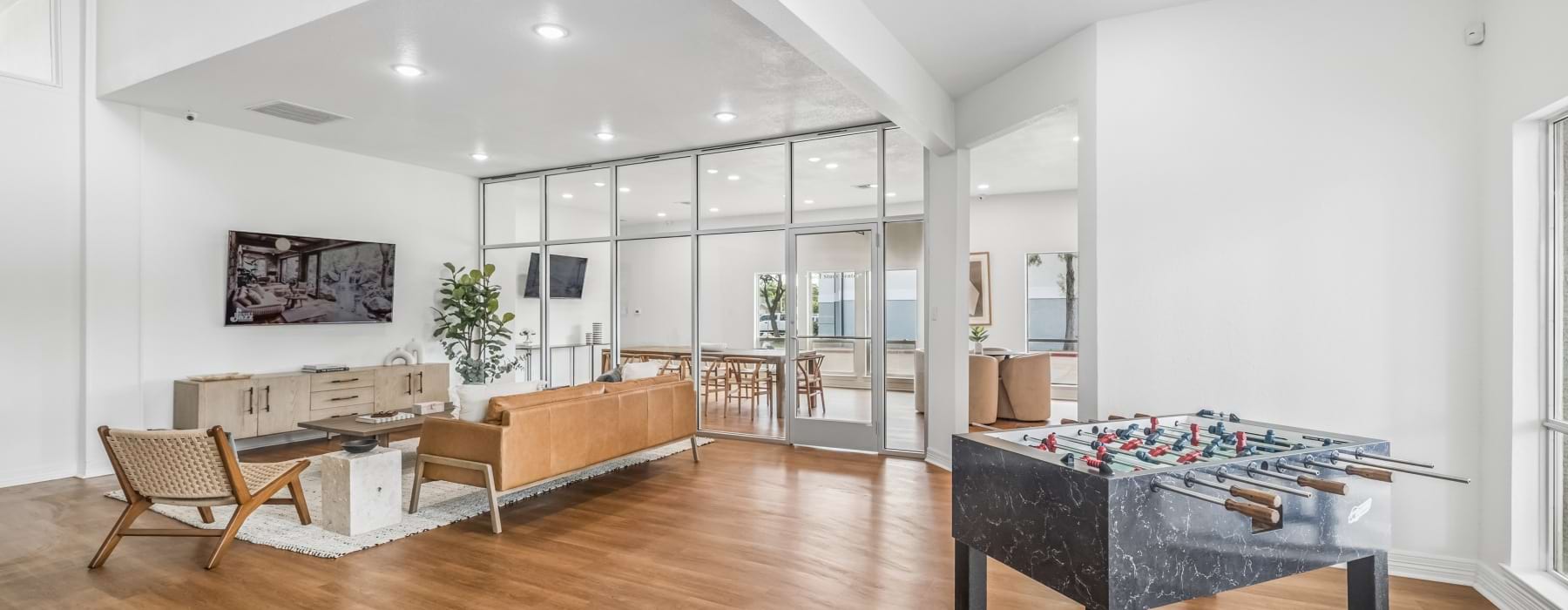 Bright, modern resident lounge featuring wood flooring, leather seating, a wall-mounted TV, foosball table, and glass-enclosed communal dining and gathering spaces.